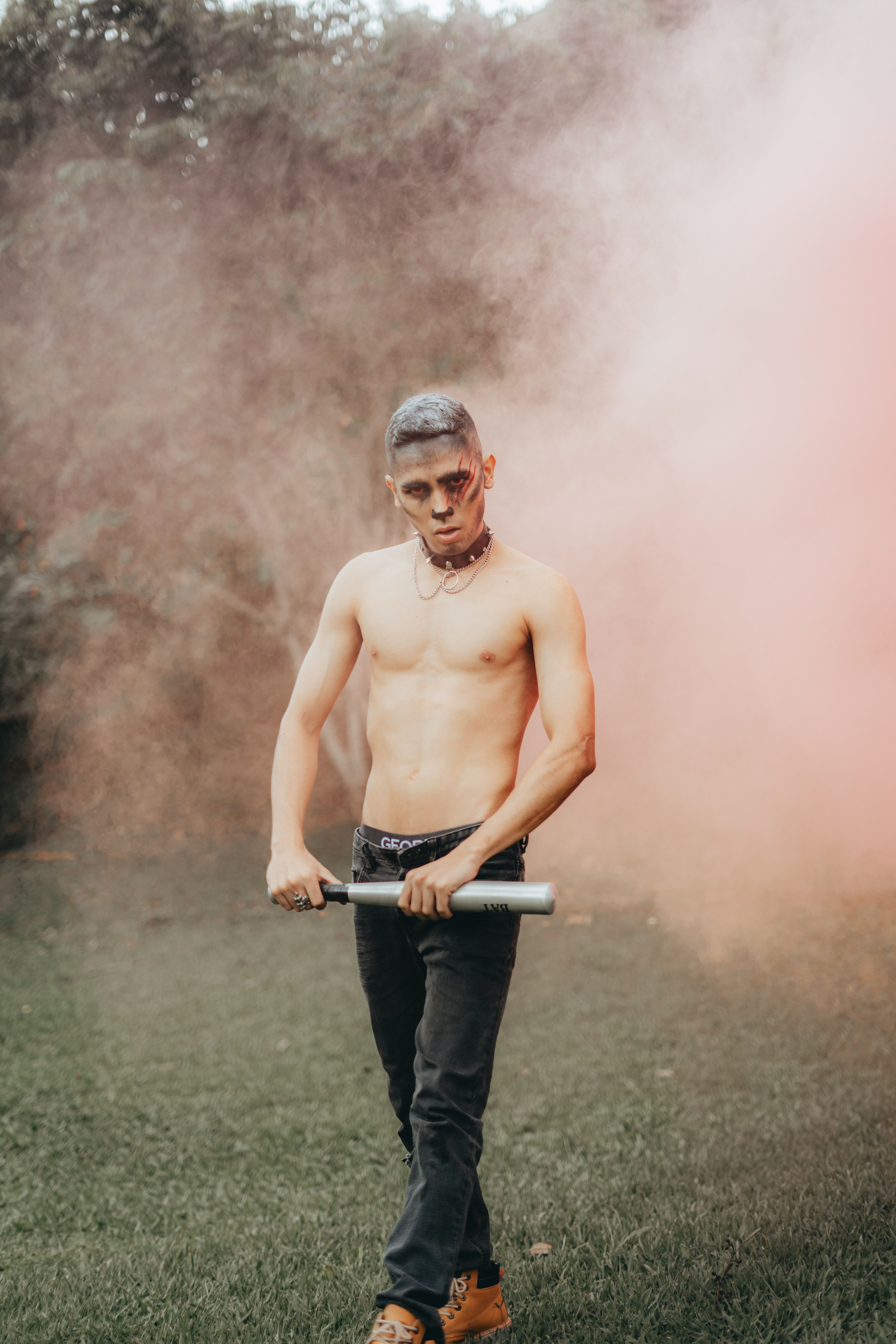 A shirtless young man stands outdoors amidst pink smoke holding a bat, exuding a moody vibe.