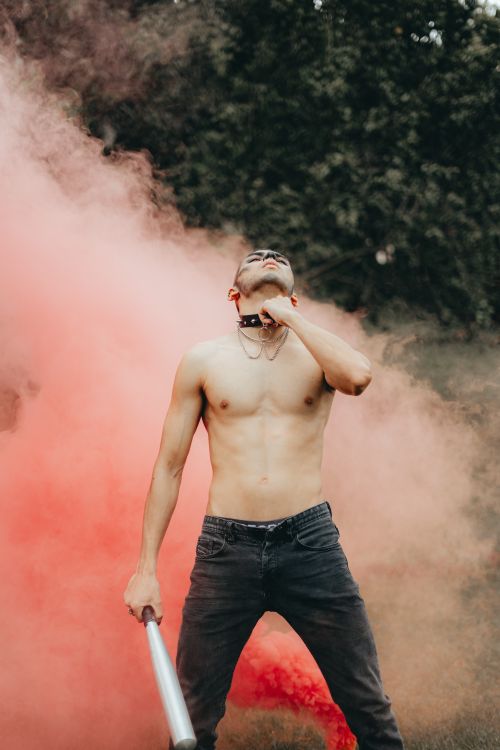 Free A shirtless man holding a bat stands confidently in red smoke outdoors, embodying performance art and creativity. Stock Photo