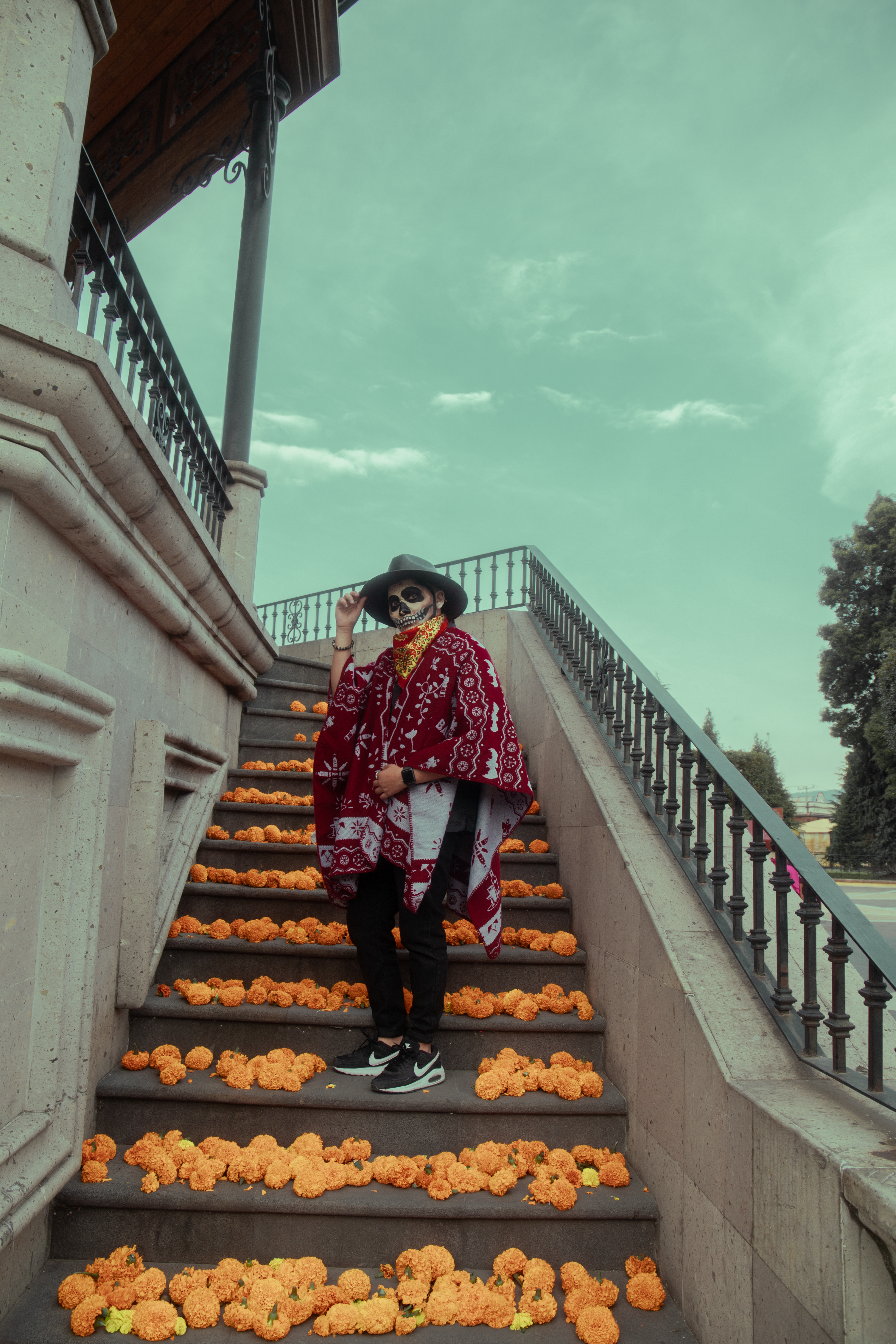 Person in traditional Day of the Dead attire stands on flower-decorated stairs outdoors.