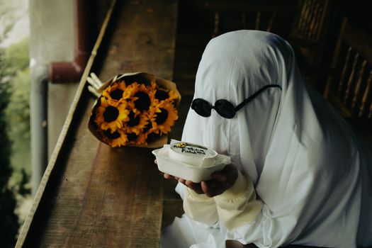 A ghostly figure in sunglasses holds a cake next to a bouquet of sunflowers. Halloween vibe in Da Lat, Vietnam.