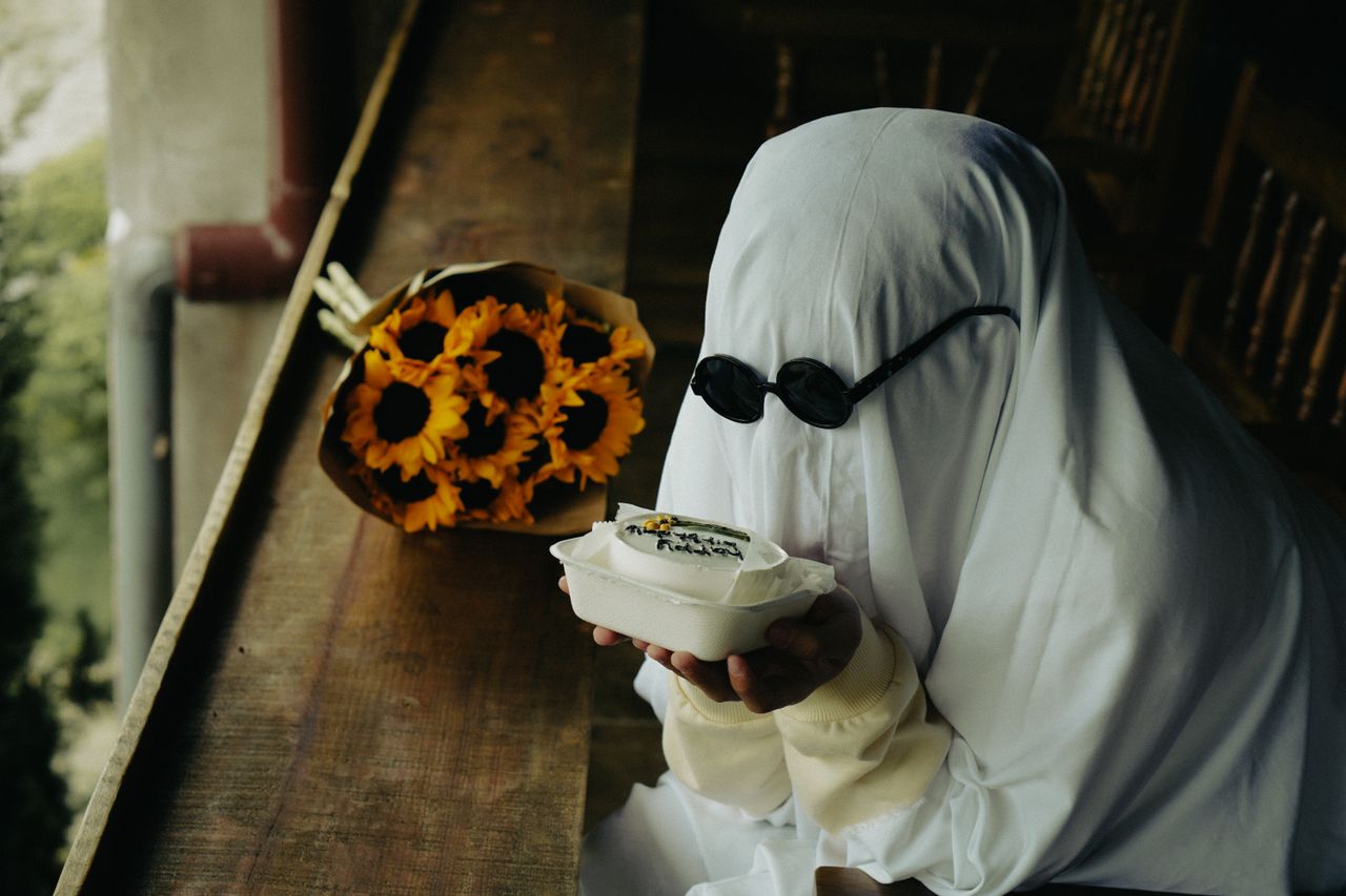 a ghostly figure in sunglasses holds a cake next to a bouquet of sunfl