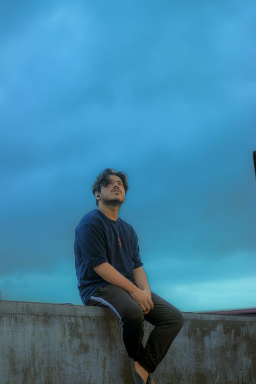 Free A man in casual attire sits on a ledge outdoors, reflecting beneath a moody, overcast sky. Stock Photo