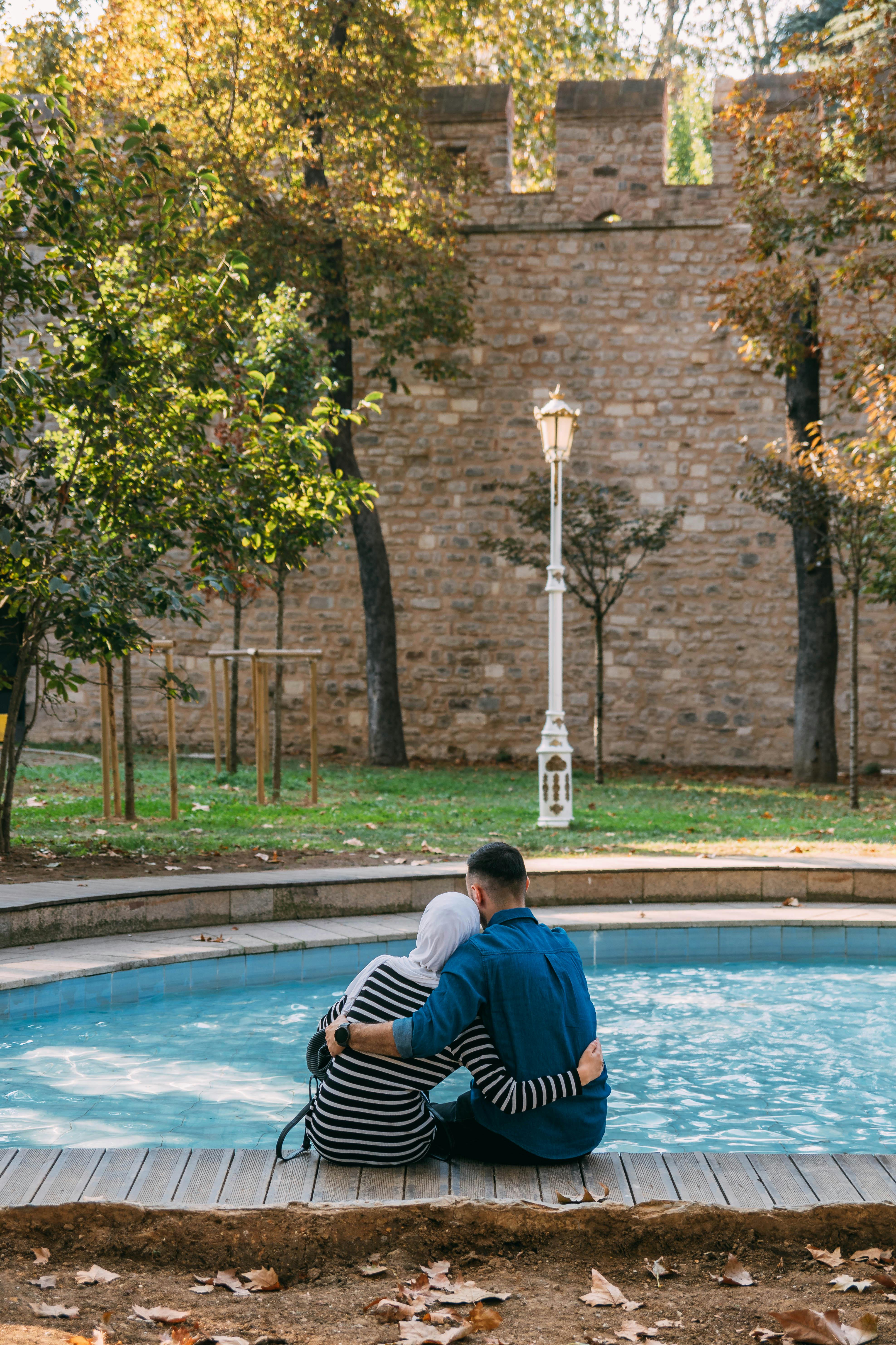 A couple embraces by a tranquil fountain in a serene park setting with autumn leaves.