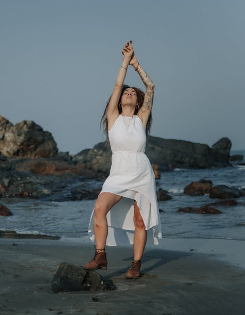 Woman in White Dress and Brown Leather Boots Standing on Beach with Raised Arms