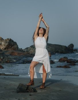Artistic fashion photography of a woman in a white dress posing with arms raised on a rocky beach.