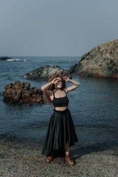 Stylish adult woman in black outfit posing by the seaside rocks under clear skies.