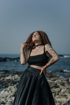 Stylish woman in a black outfit poses confidently by rocky ocean shore.