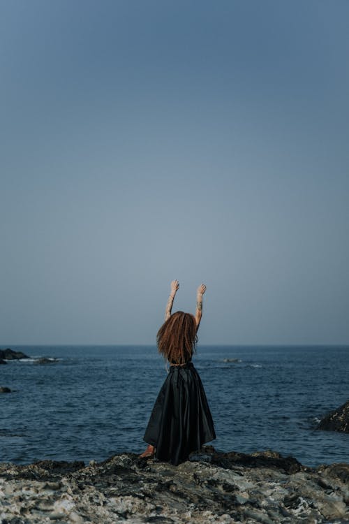 Back View of a Woman in a Black Dress Standing on a Shore with Arms Raised 