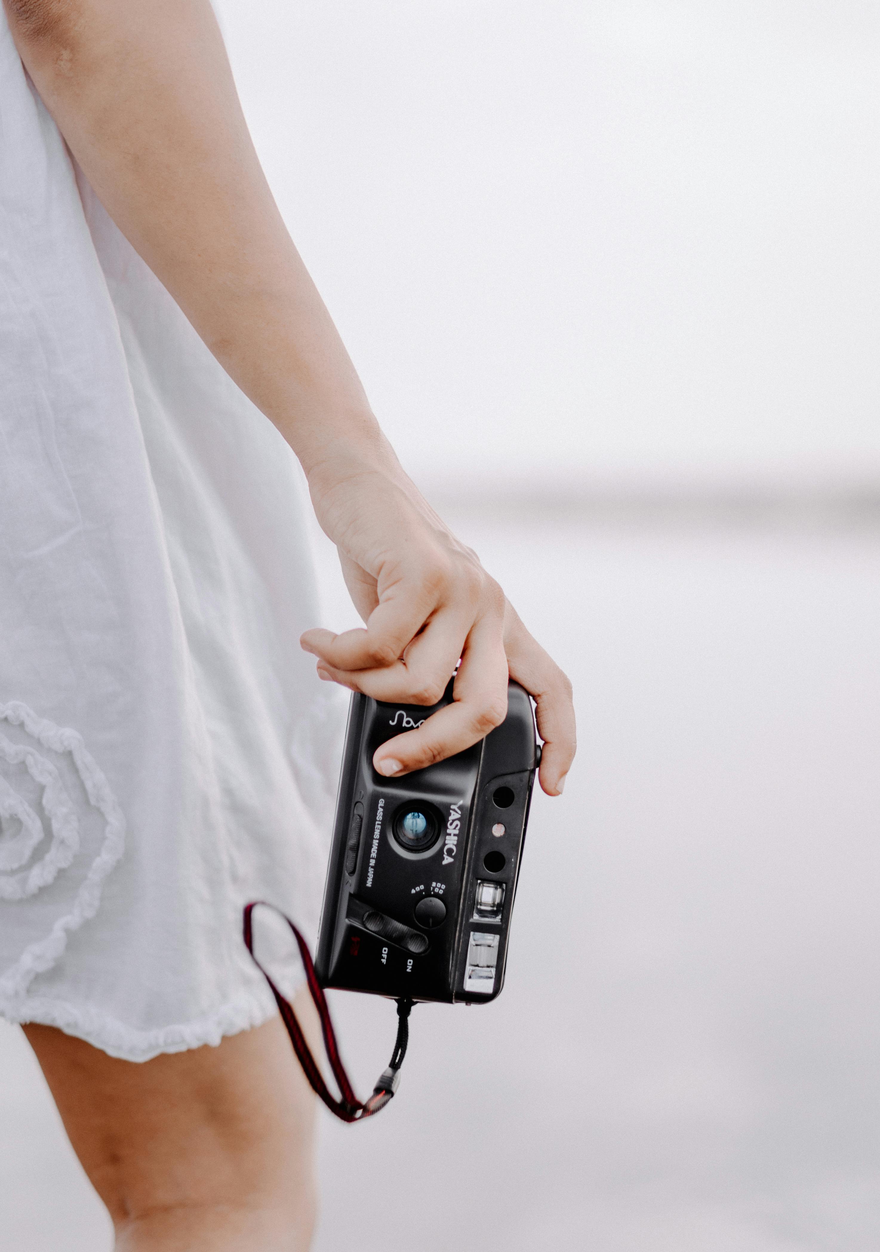A woman holds a classic analog camera, highlighted by a white dress in an outdoor setting.