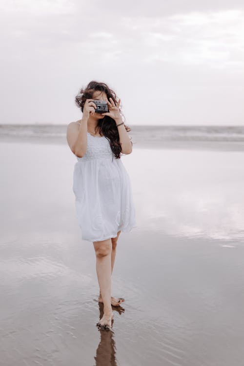 Woman in White Dress Taking Pictures with Camera on Sea Shore