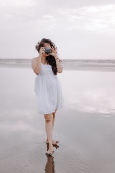A woman in a white dress taking pictures on a serene beach shore.