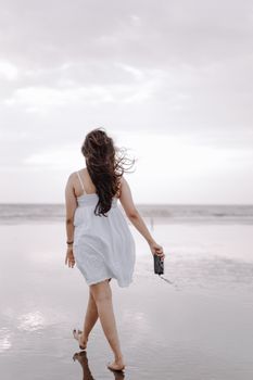 Woman in white dress walking on beach holding a camera, calm seaside setting.