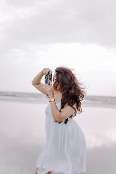Elegant woman in a white dress photographs the serene beach, capturing the sea's beauty.