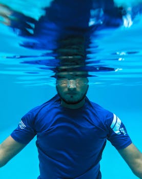 A man swimming underwater in a clear blue pool with reflections at the surface.