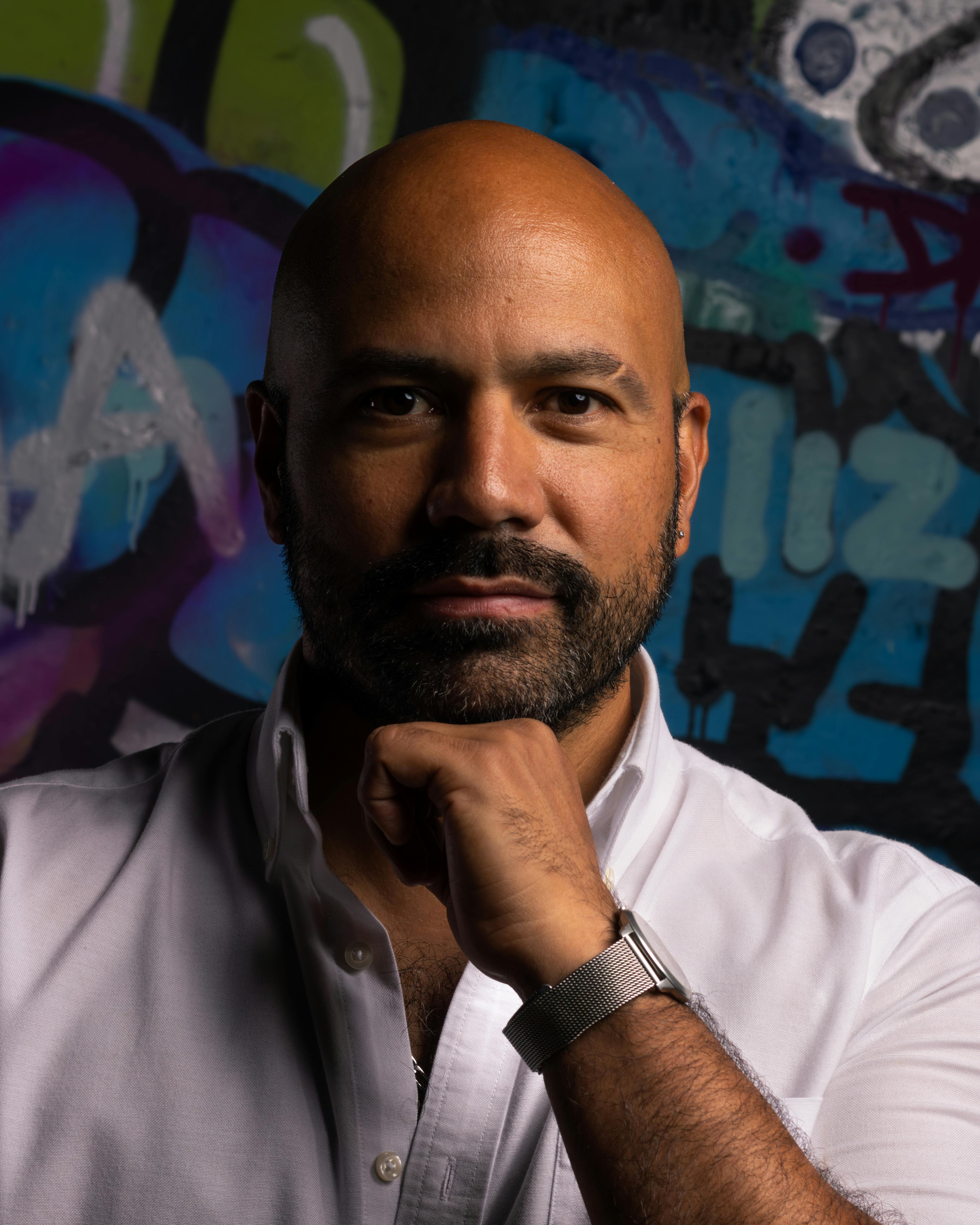 Bald man in a white shirt posing thoughtfully in front of a colorful graffiti wall.