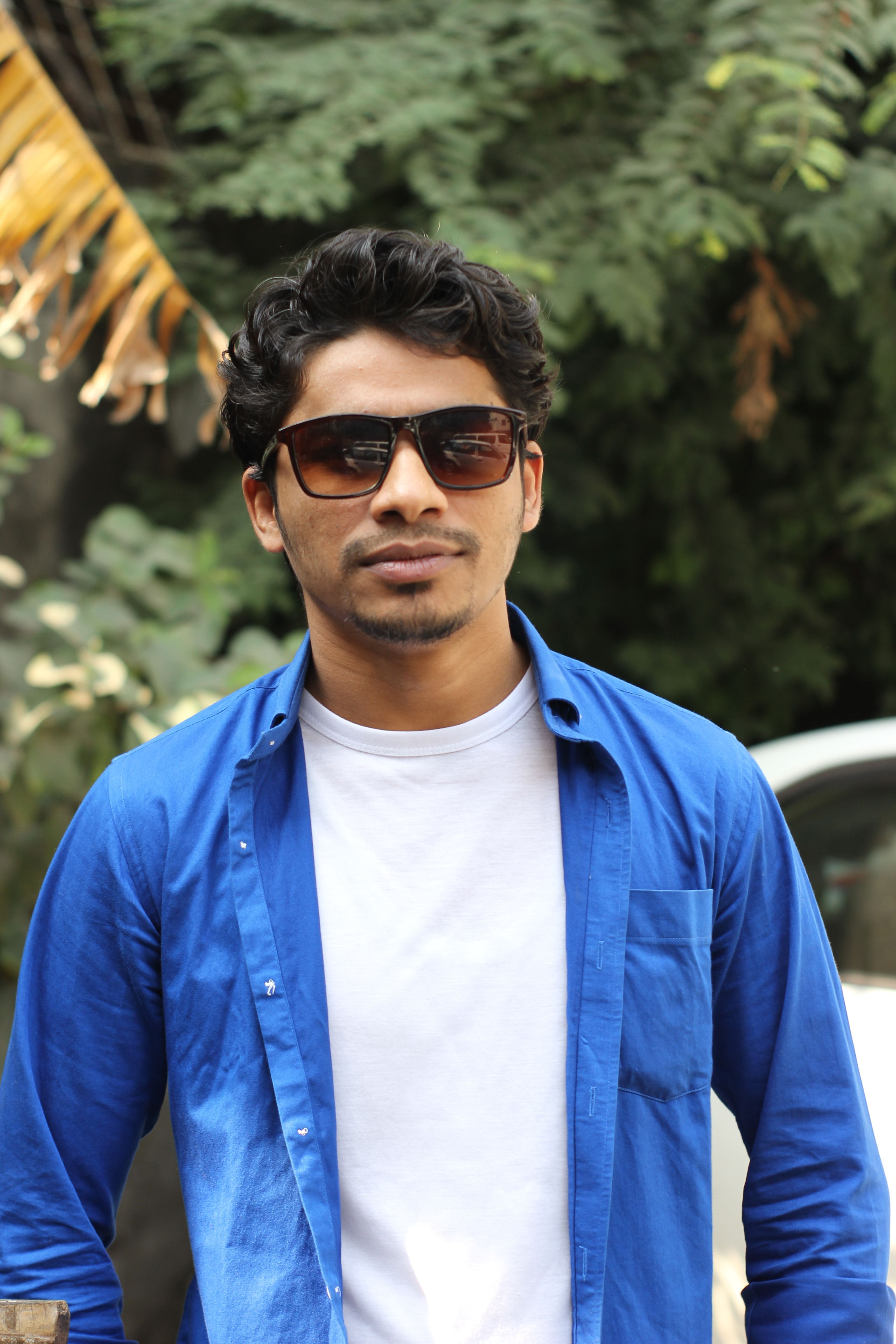 Portrait of a young man wearing sunglasses and a blue shirt, outdoors with a natural backdrop.