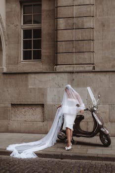 Elegant bride in wedding dress poses stylishly with a scooter on city streets.