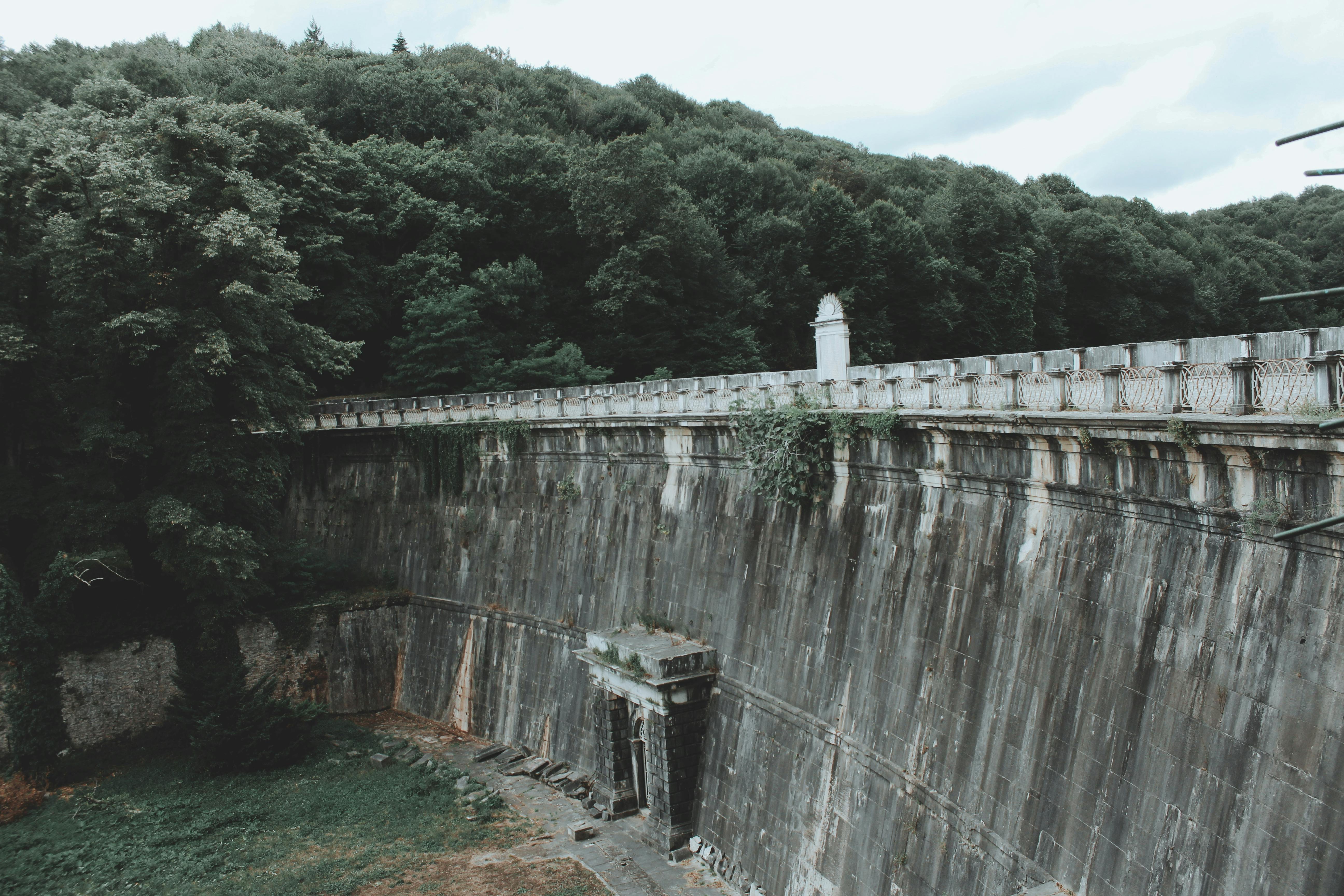 A vintage stone dam stands tall beside a dense forest under an overcast sky, creating a moody scene.