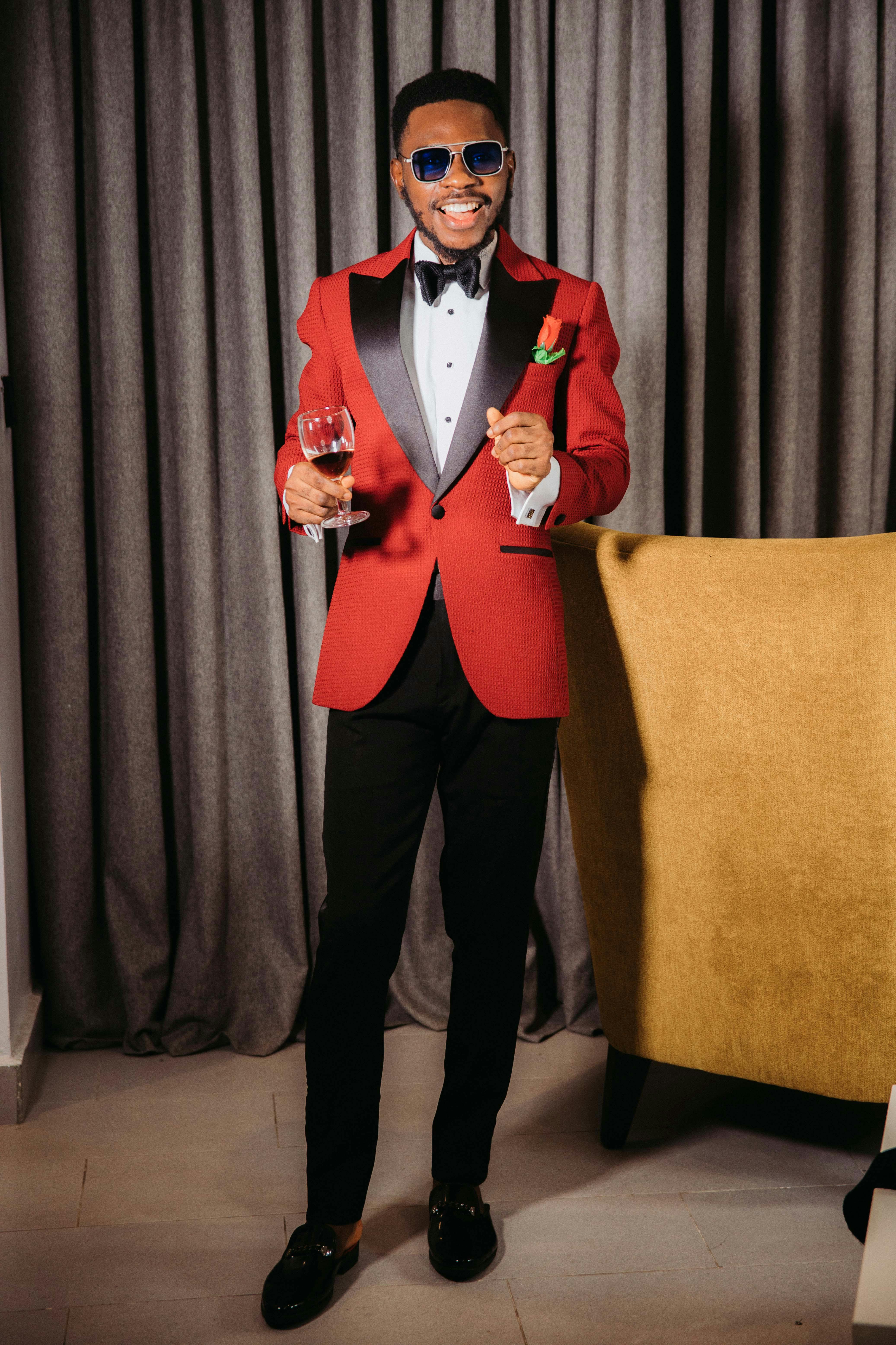 A stylish man in a red suit with sunglasses standing indoors holding a wine glass, exuding elegance and confidence.
