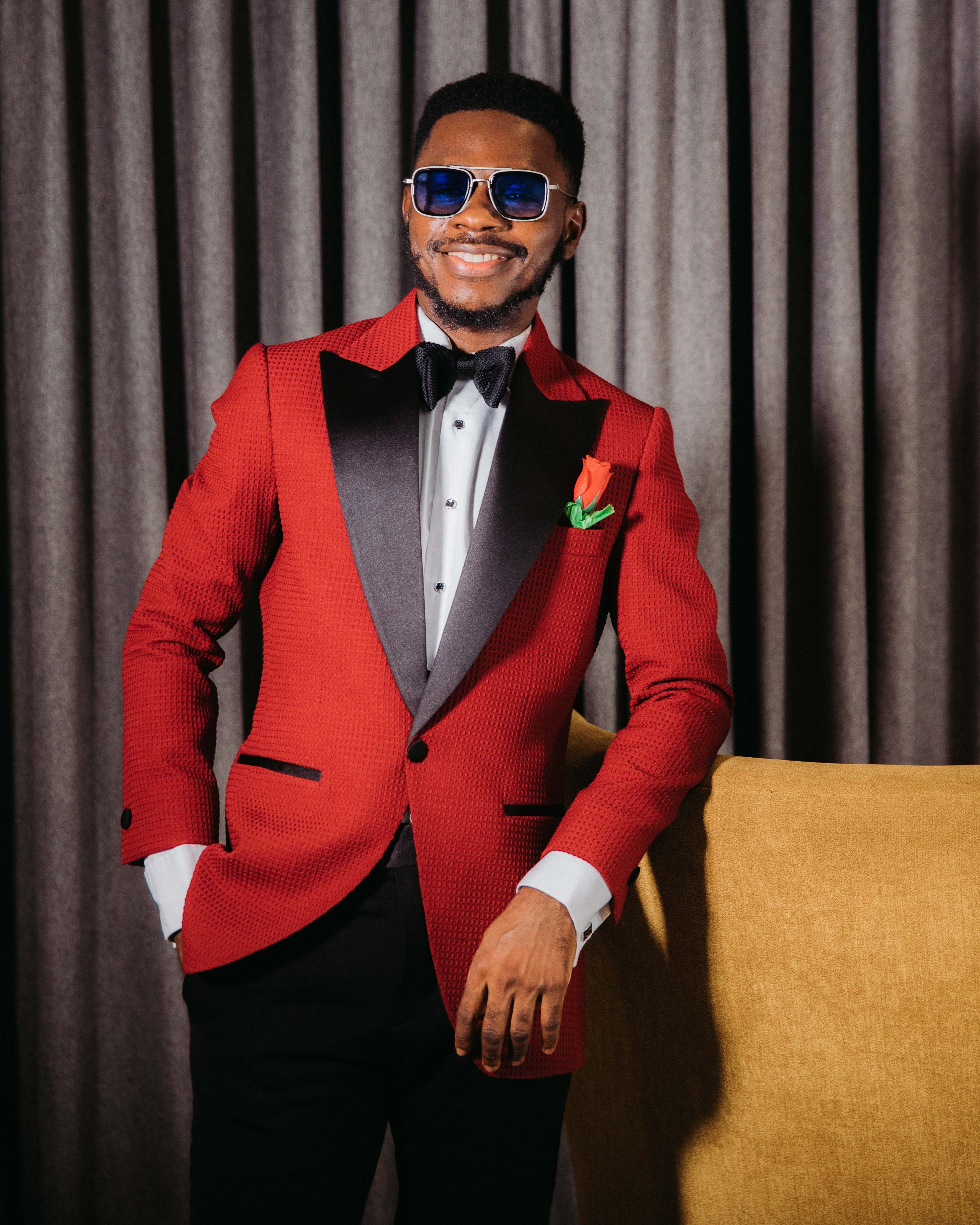 Elegant fashion portrait of a man in a red tuxedo and sunglasses, smiling indoors.