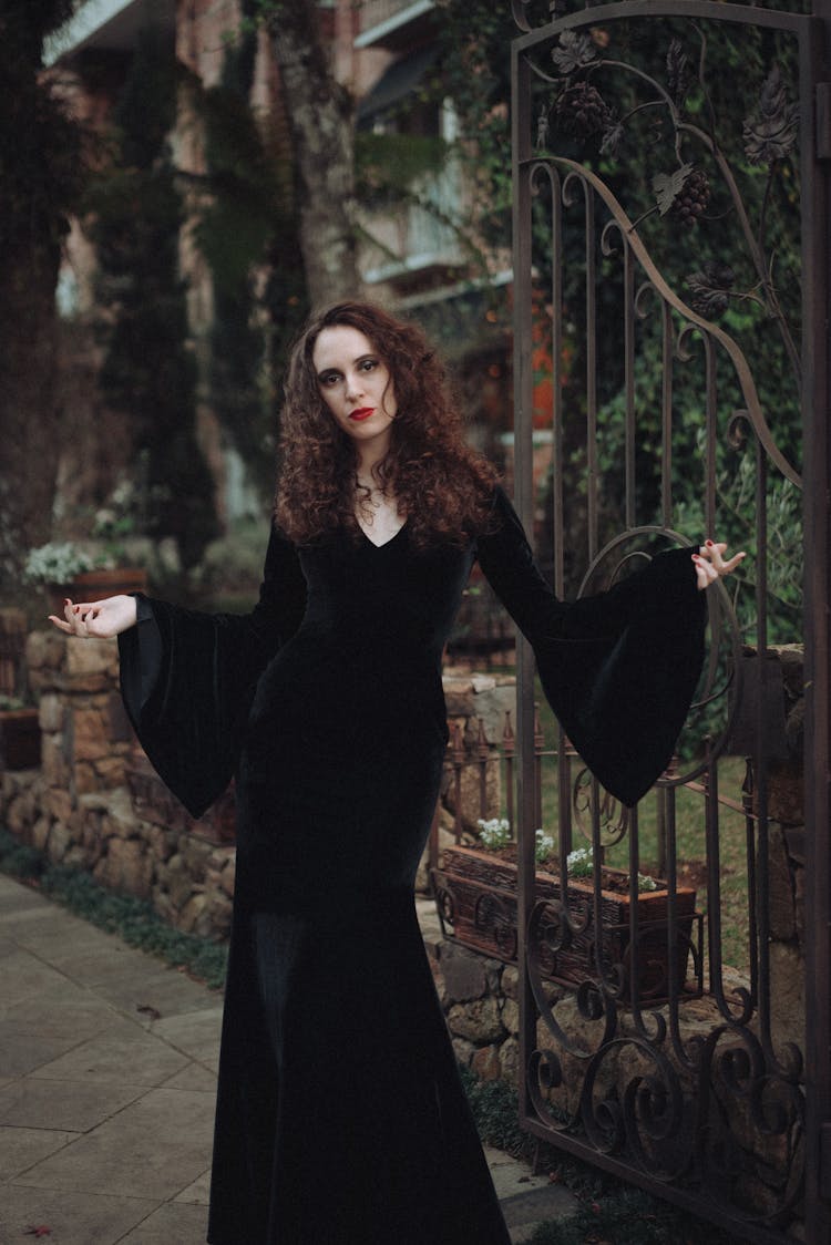 Woman In Black Gown Standing By Open Iron Gate