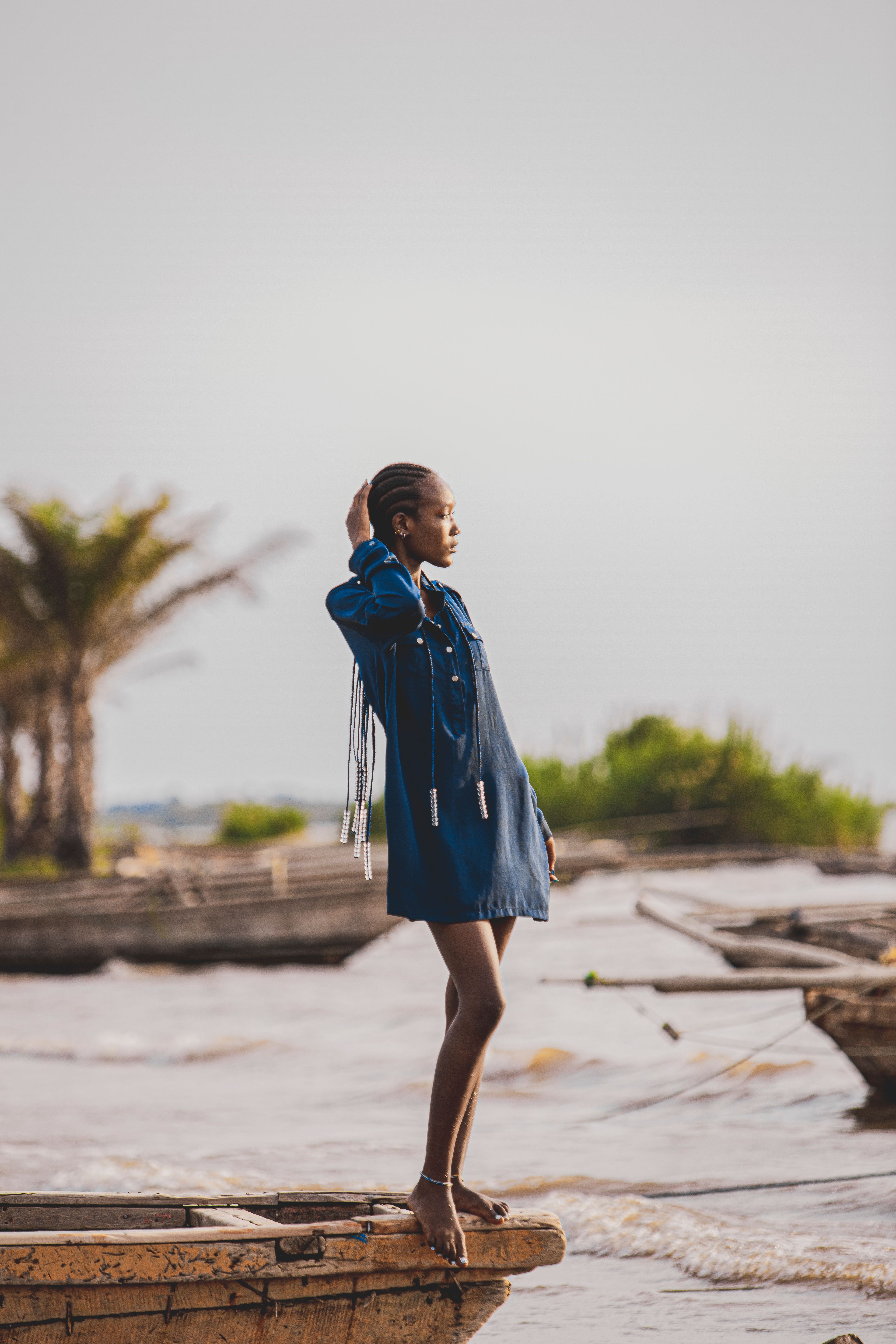 Black woman in a fashionable dress elegantly stands on a wooden boat during a sunset by the water.