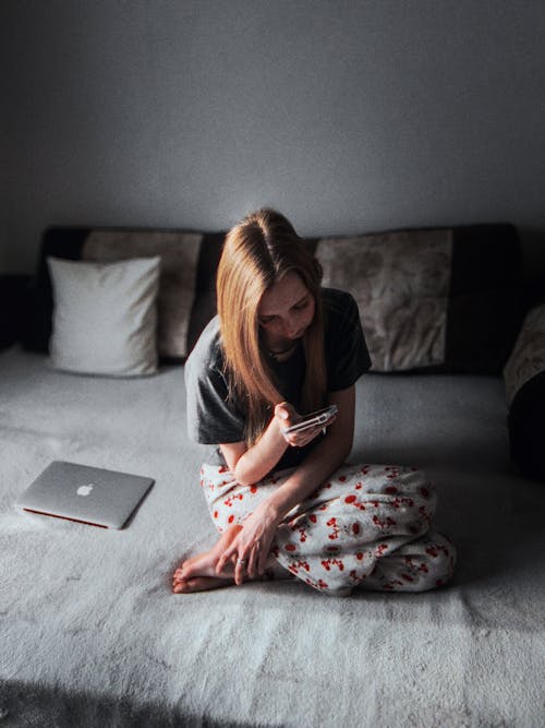 Free Young Woman in Pajamas Sitting on the Bed with a Phone in Her Hand Stock Photo