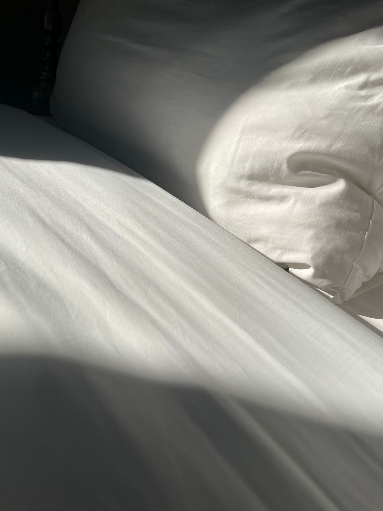 White pillow and silky white bedsheet with sunlight casting shadows across the bed