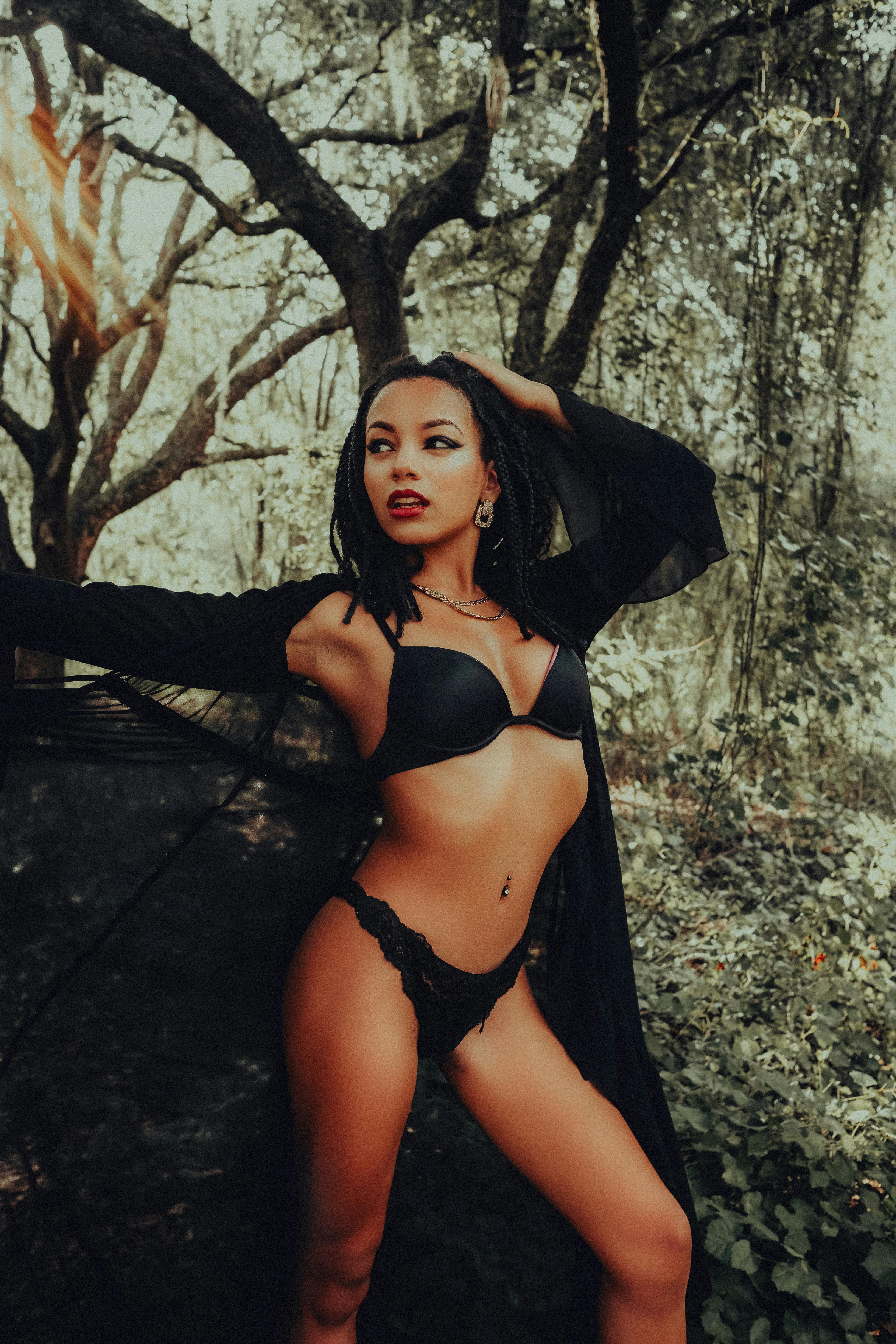 Elegant woman posing in black lingerie and cloak amidst a lush forest background.