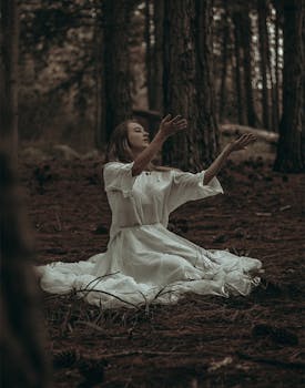 A woman in a white dress sits in a forest with arms raised, evoking mystery.
