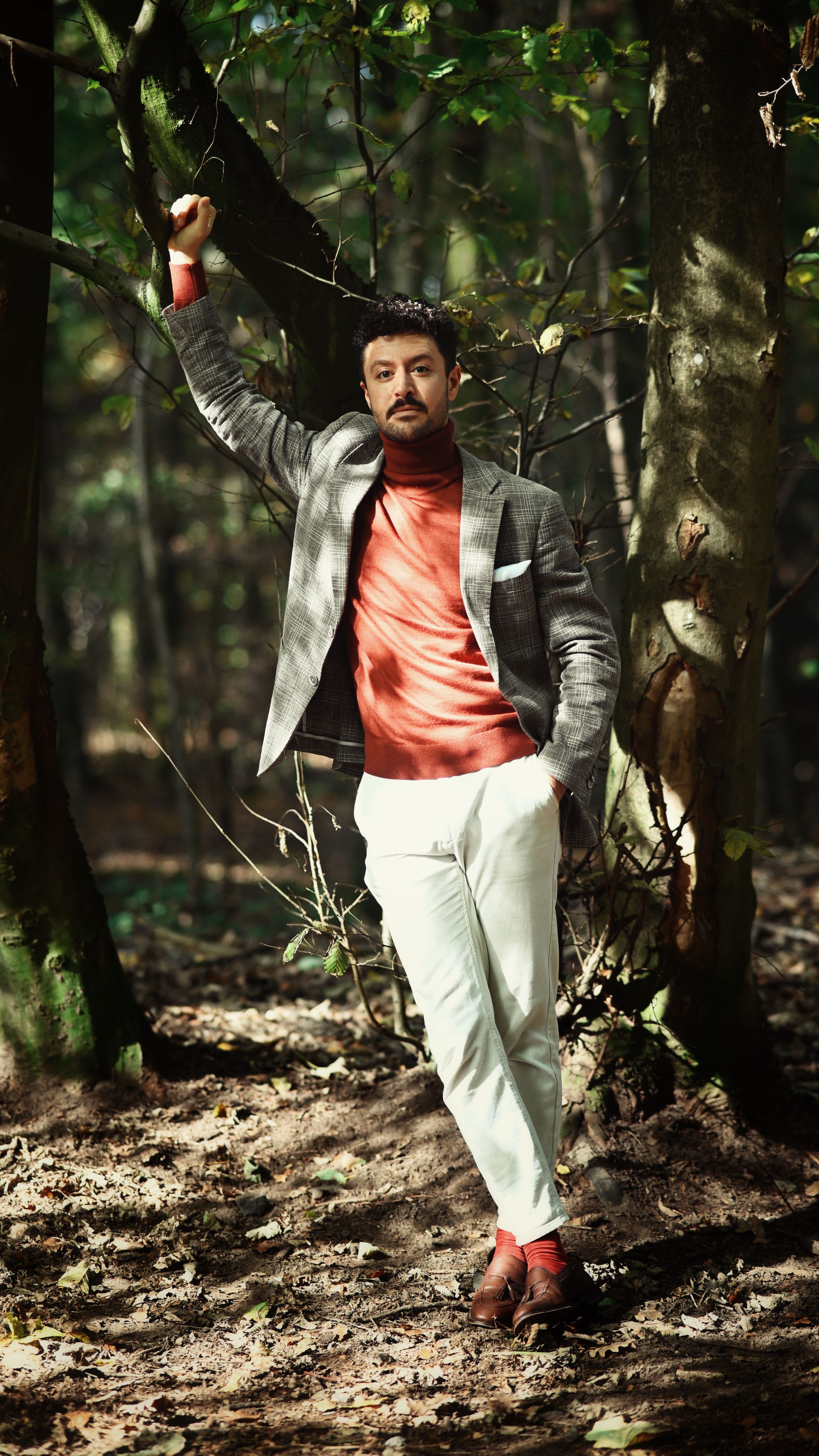 Elegant man in a blazer and turtleneck stands in a forest, exuding style and confidence.