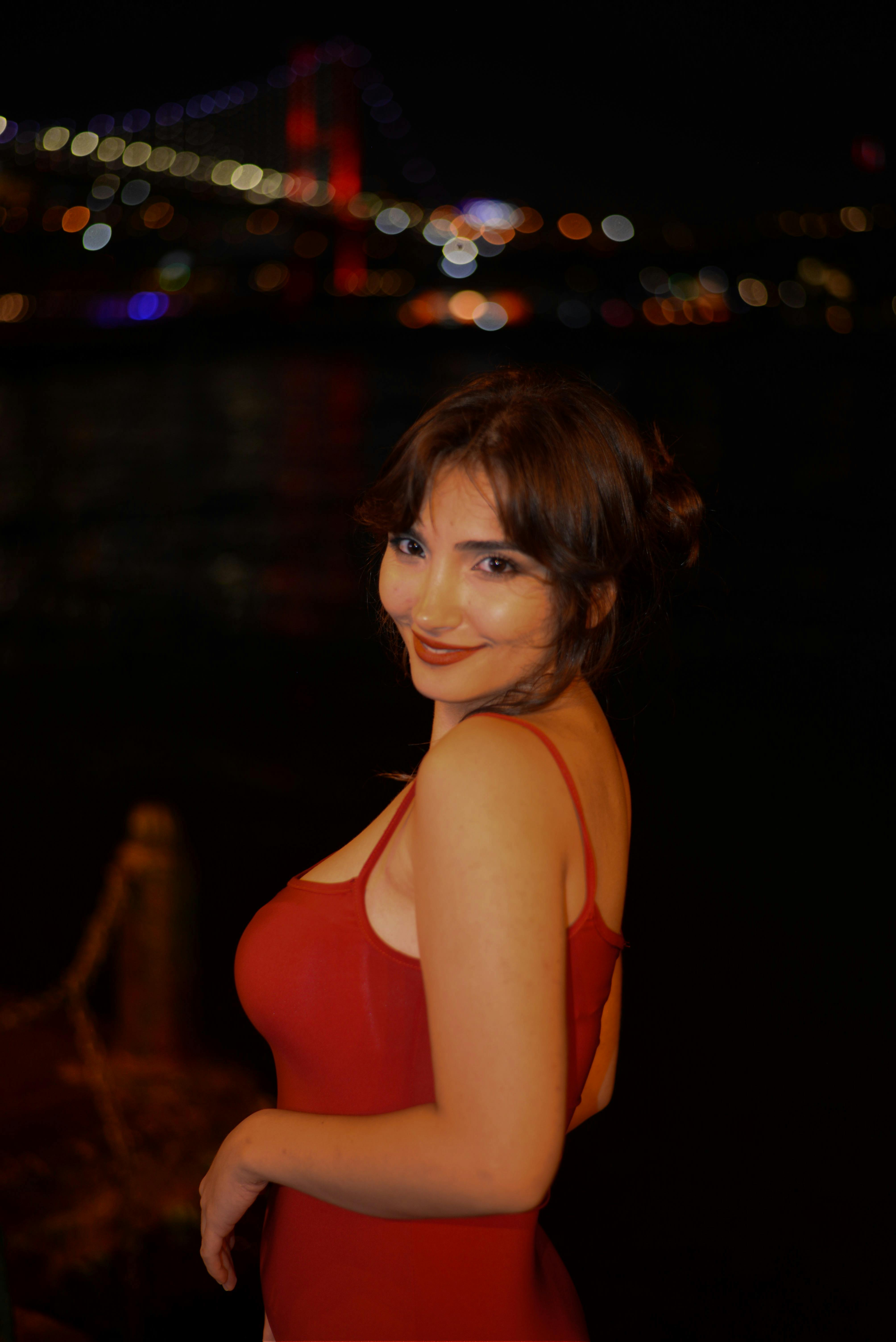 Young woman in red dress posing by a waterfront at night with city lights in the background.