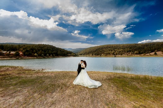 A newlywed couple shares a romantic embrace on lakeside, surrounded by nature and a vibrant blue sky.