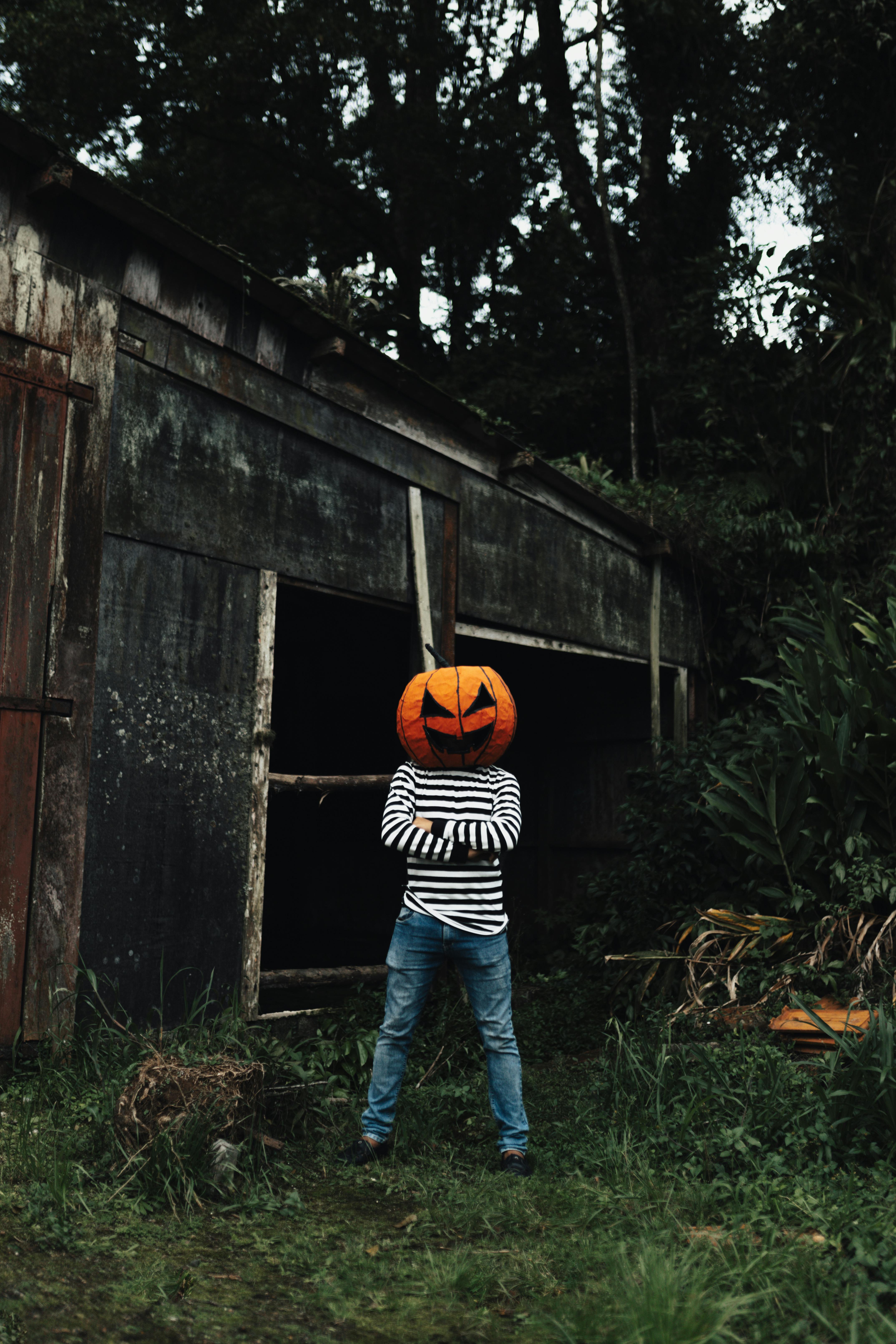 Person with a pumpkin head costume standing with arms crossed by an old shed