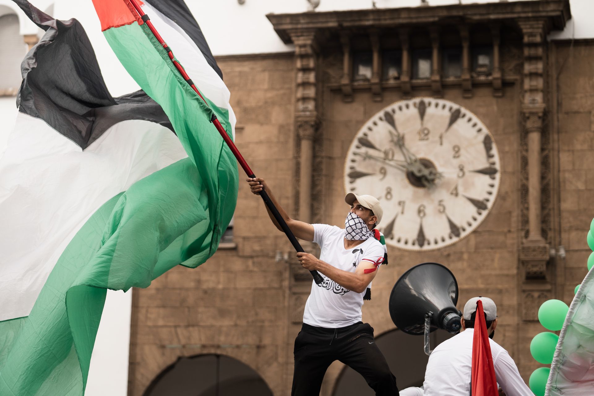 Protester passionately waves Palestinian flag during a demonstration in front of a historic clock tower.