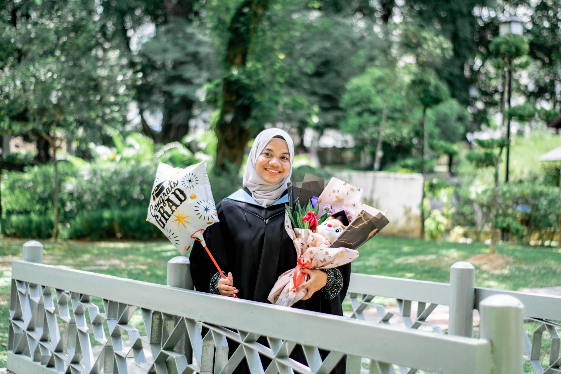 Smiling Muslim woman in hijab holding graduation balloon and flowers outdoors in park.