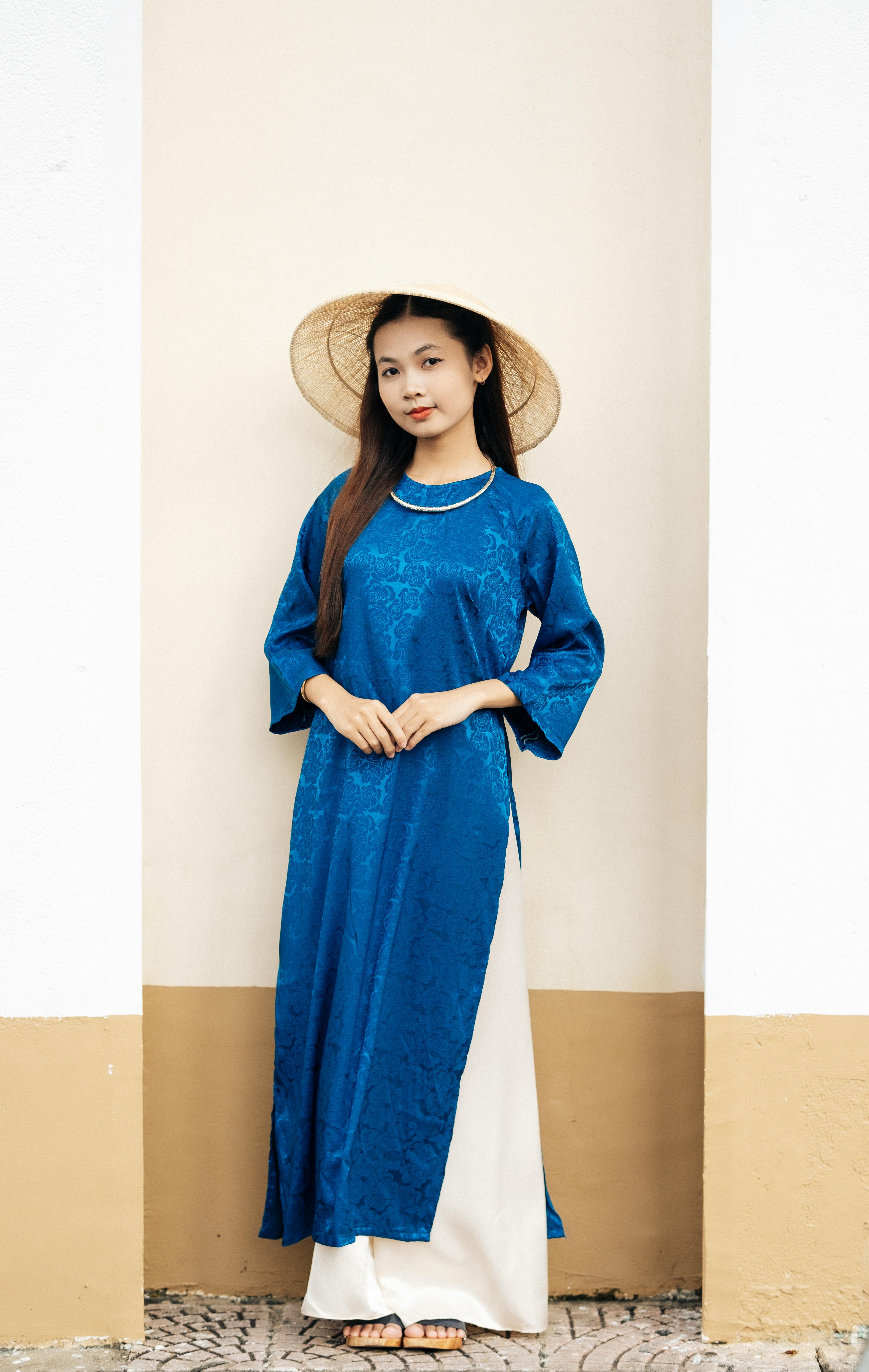 Young Asian woman wearing a traditional blue gown and conical hat, standing against a wall outdoors.