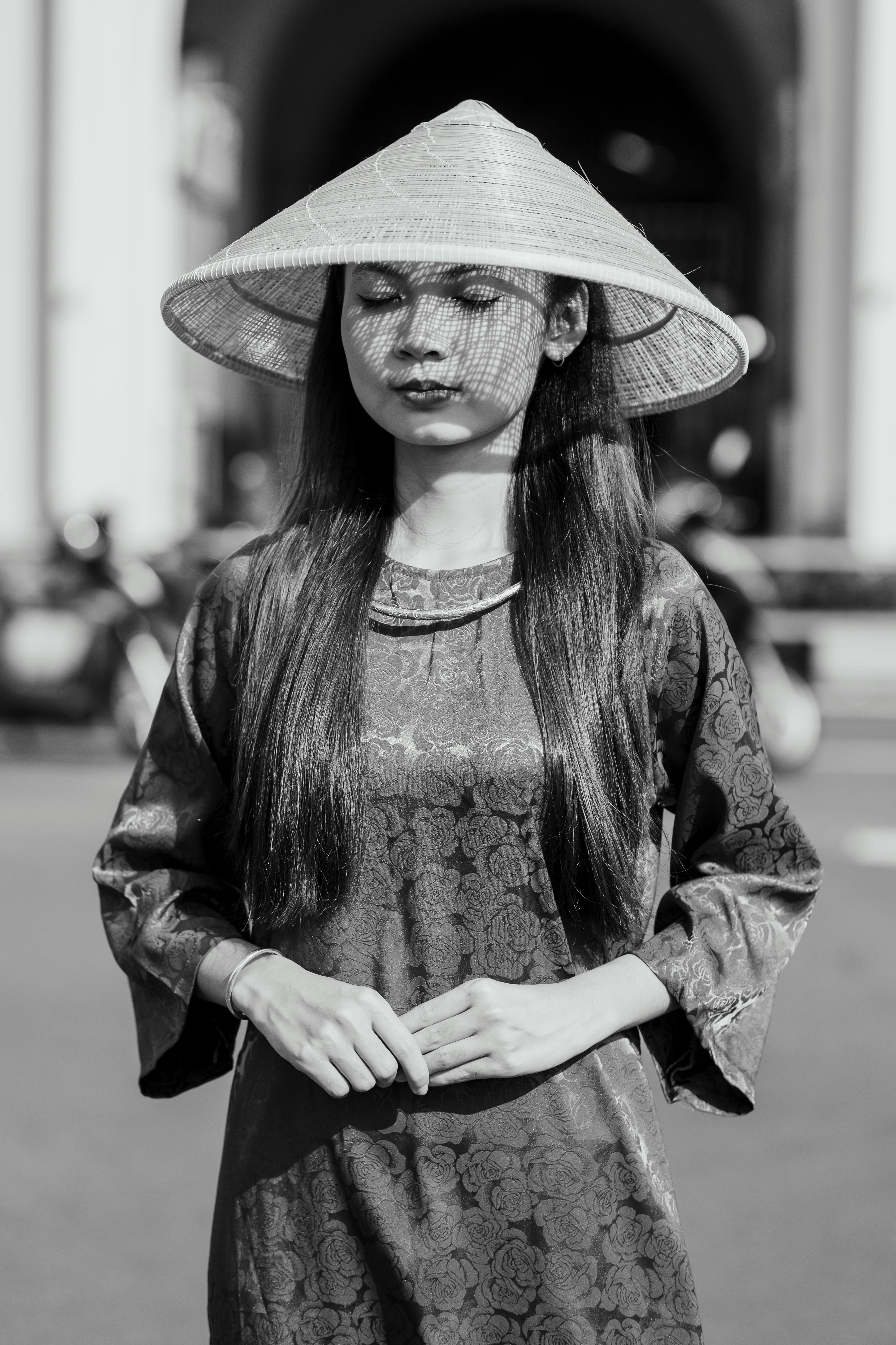 Young woman in traditional conical hat standing outdoors with eyes closed in a serene pose.