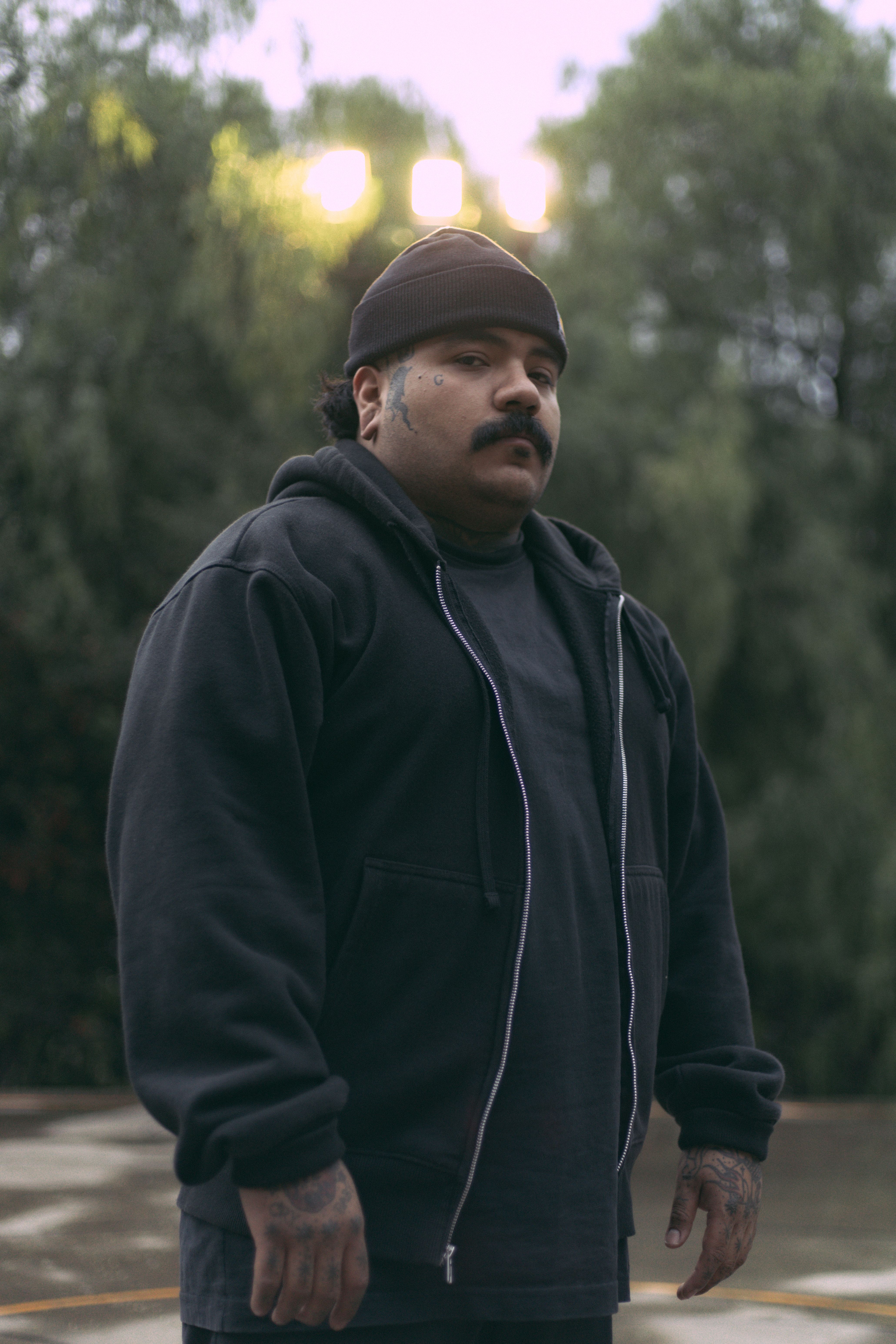 Portrait of a man in a hoodie with a mustache and tattoos posing outdoors.