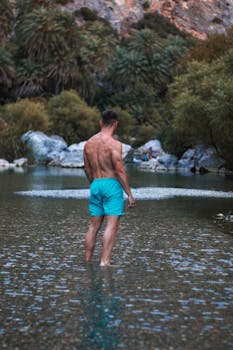A man in turquoise shorts stands shirtless in a tropical river, surrounded by lush greenery.