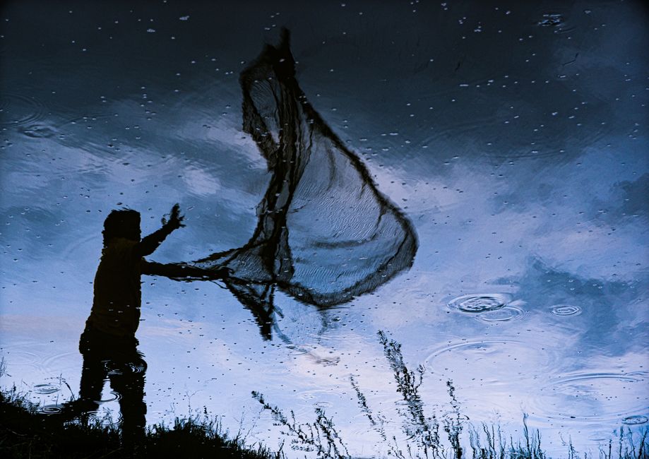Silhouette of a fisherman casting a net reflected in a tranquil lake in Wayanad, India.