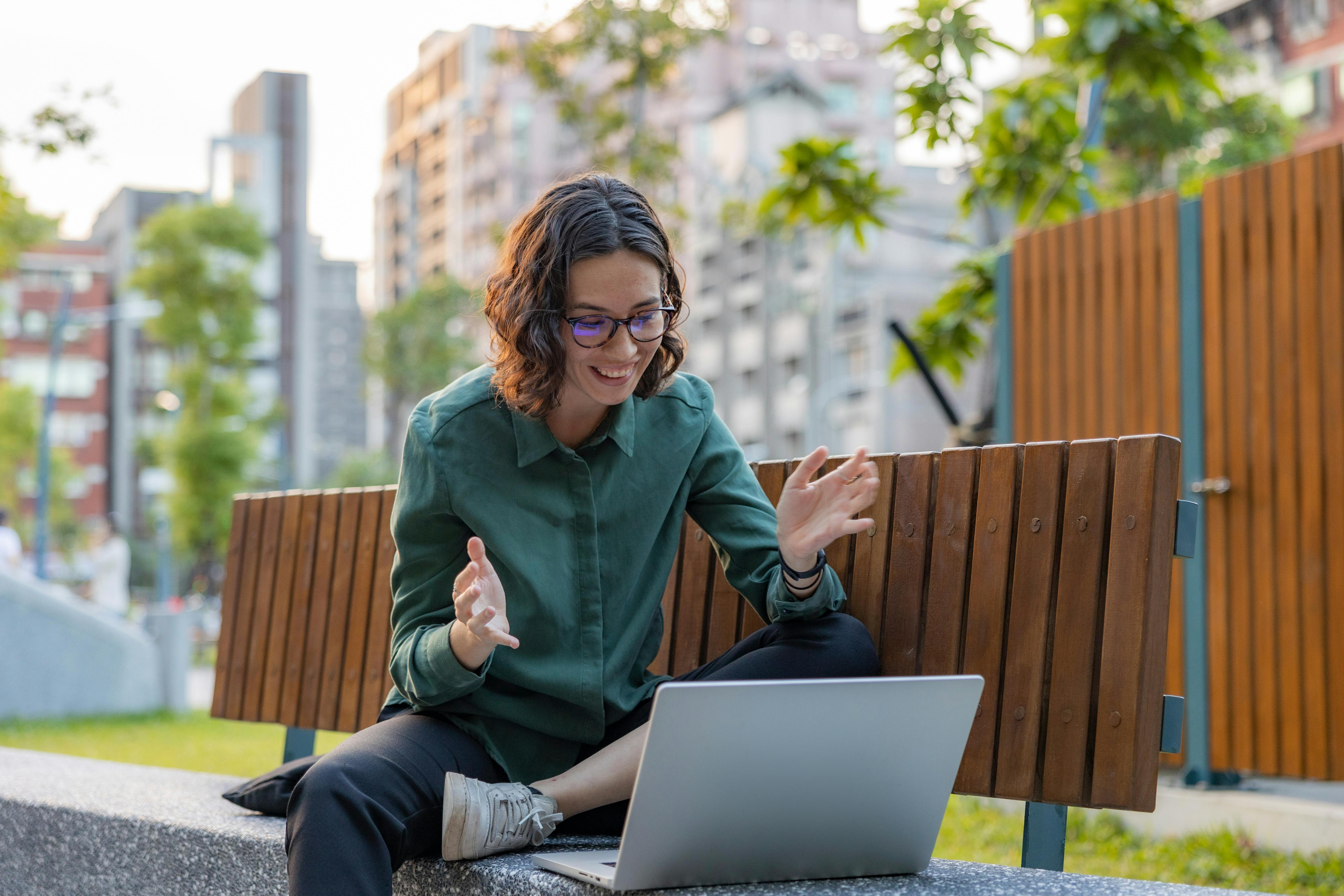 young woman smiling and doing remote work video call at park bench