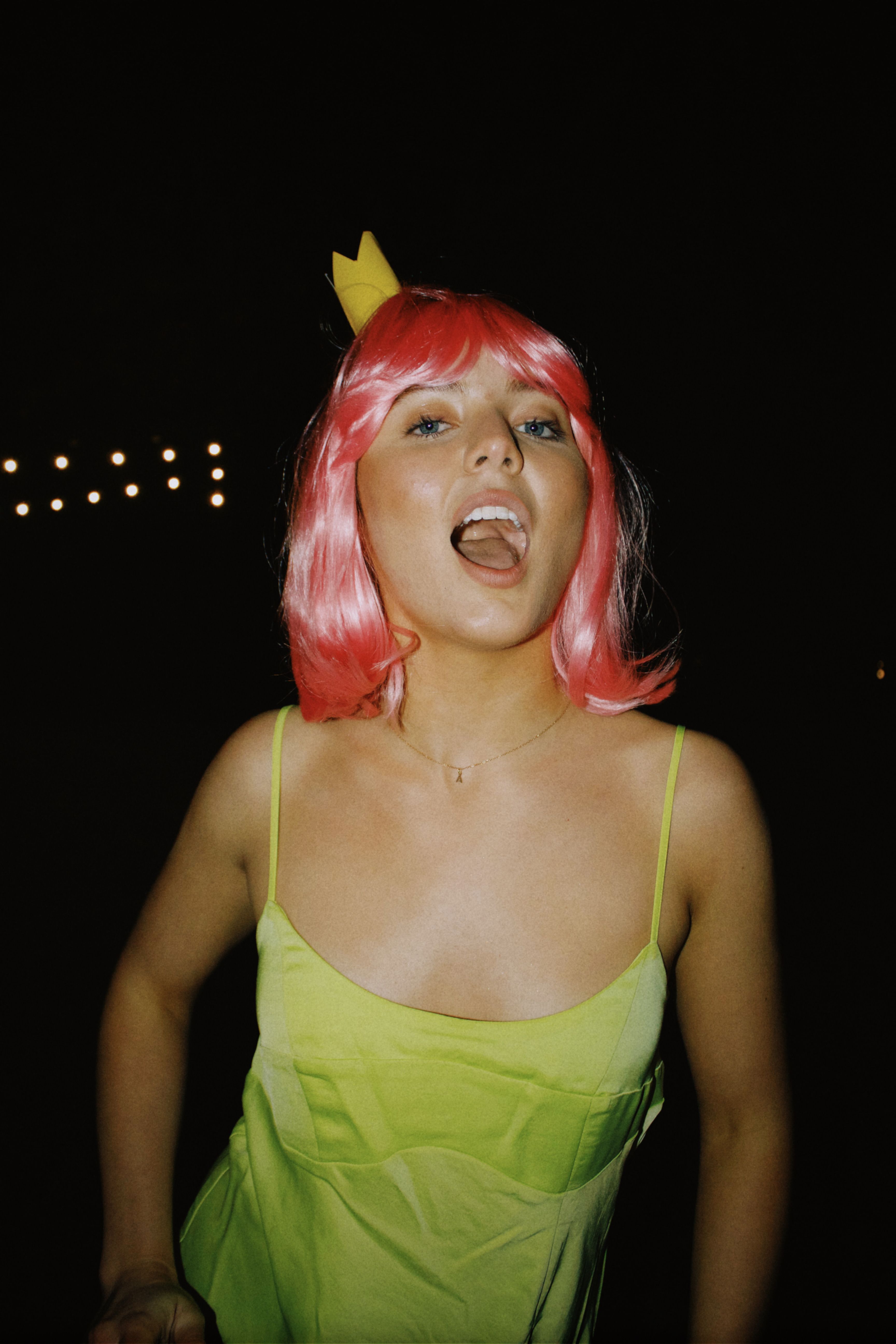 Young woman with pink hair and a yellow dress celebrating at night outdoors.