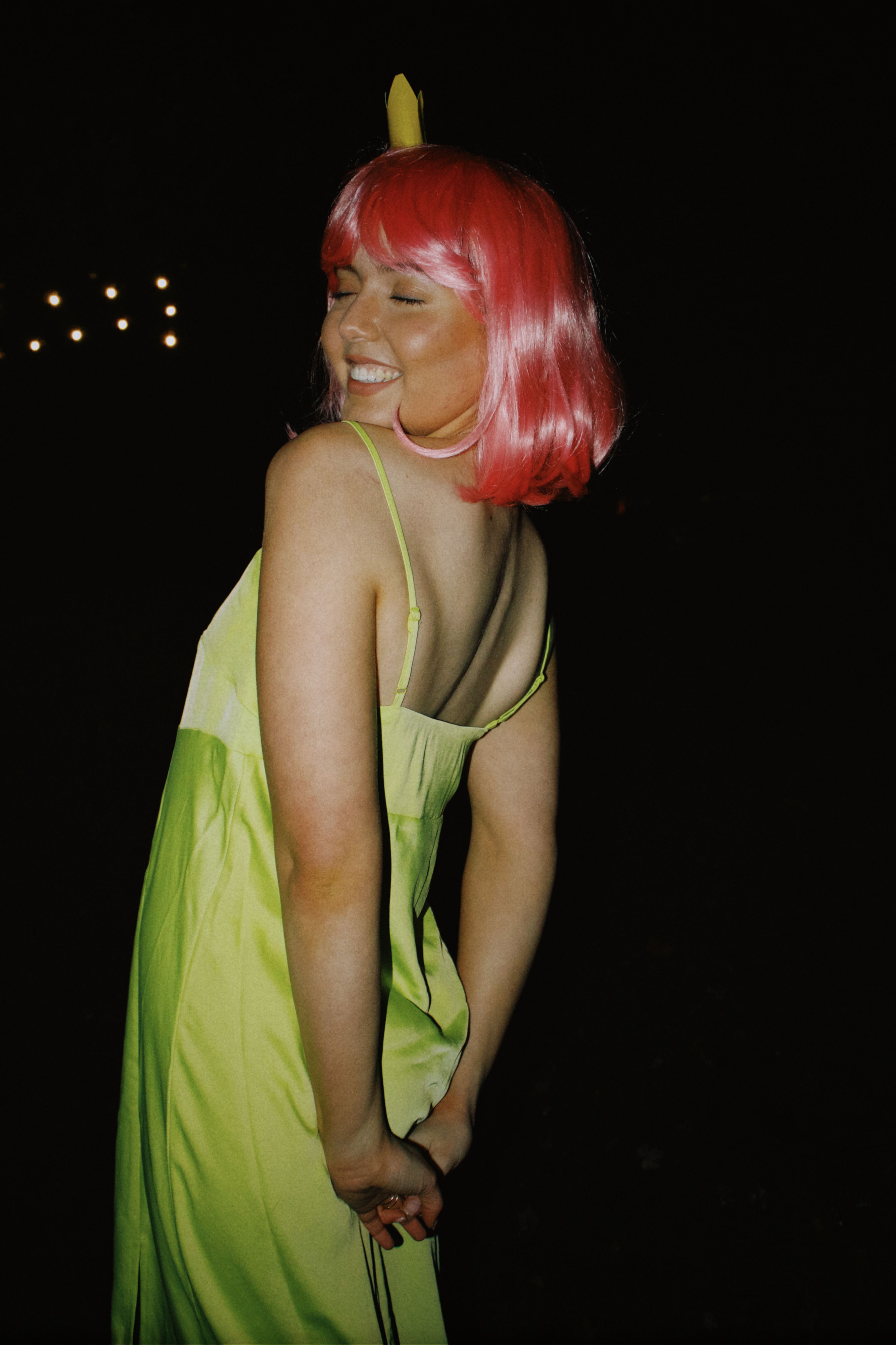 A joyful woman with pink hair in a green dress, posing playfully at night.