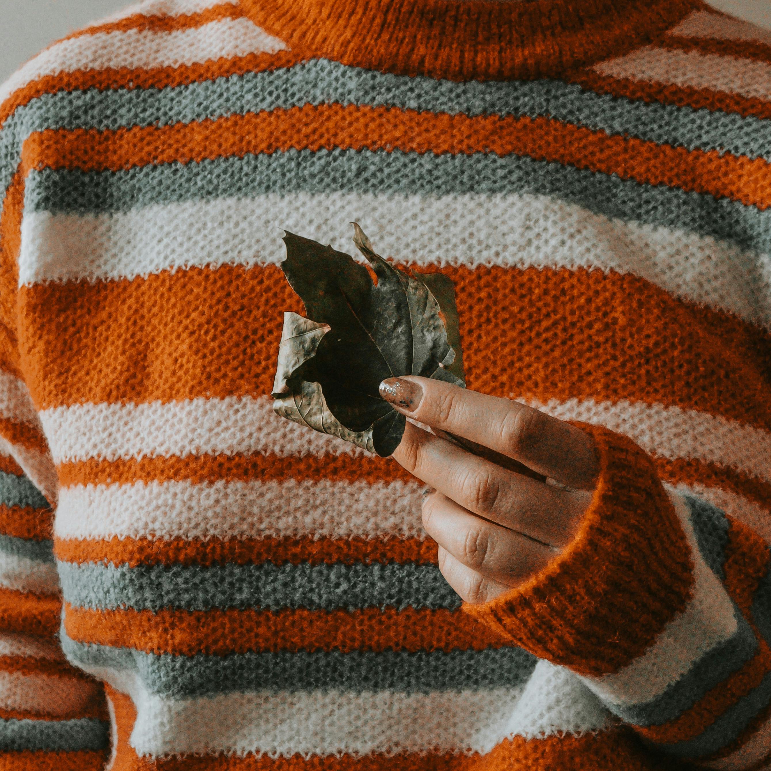 Hand holding a dry leaf against a colorful sweater background, symbolizing autumn warmth.