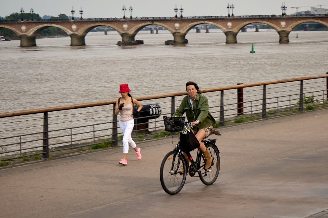 Bordeaux, France, 05.23.2023 A cycling woman in trendy attire blends harmoniously with urban life. Pont de Pierre (Stone Bridge) stretches over the tranquil water of the river Garonne.