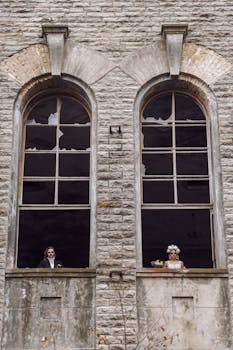 A couple in Halloween costumes and makeup in an abandoned building's windows creating a spooky scene.