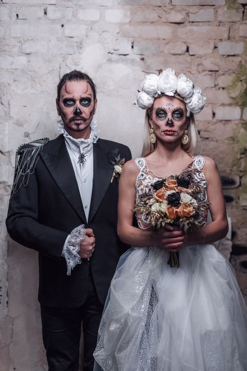 Free A couple in Santa Muerte makeup and wedding attire standing in front of a rustic wall. Stock Photo