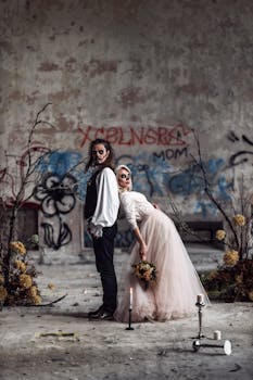 A couple stands in Santa Muerte costumes, holding a bouquet, surrounded by candles and graffiti, embracing Halloween spirit.