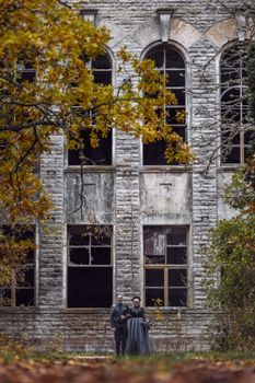 A mysterious couple dressed in dark costumes outside an abandoned building with autumn foliage.